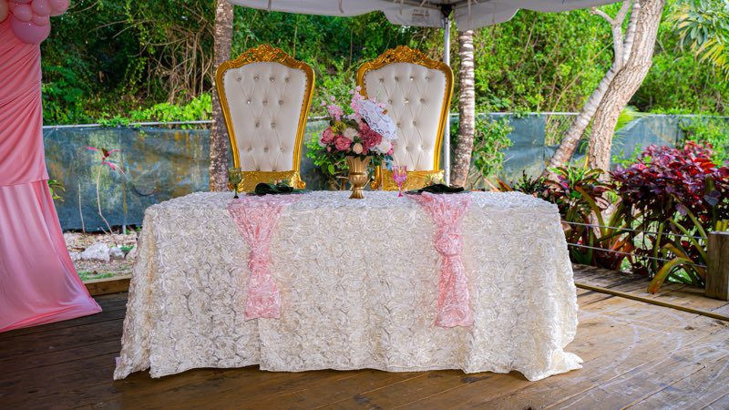 Two white and gold throne chairs behind a white rosette tablecloth, pink sashes and a floral centrepiece.
