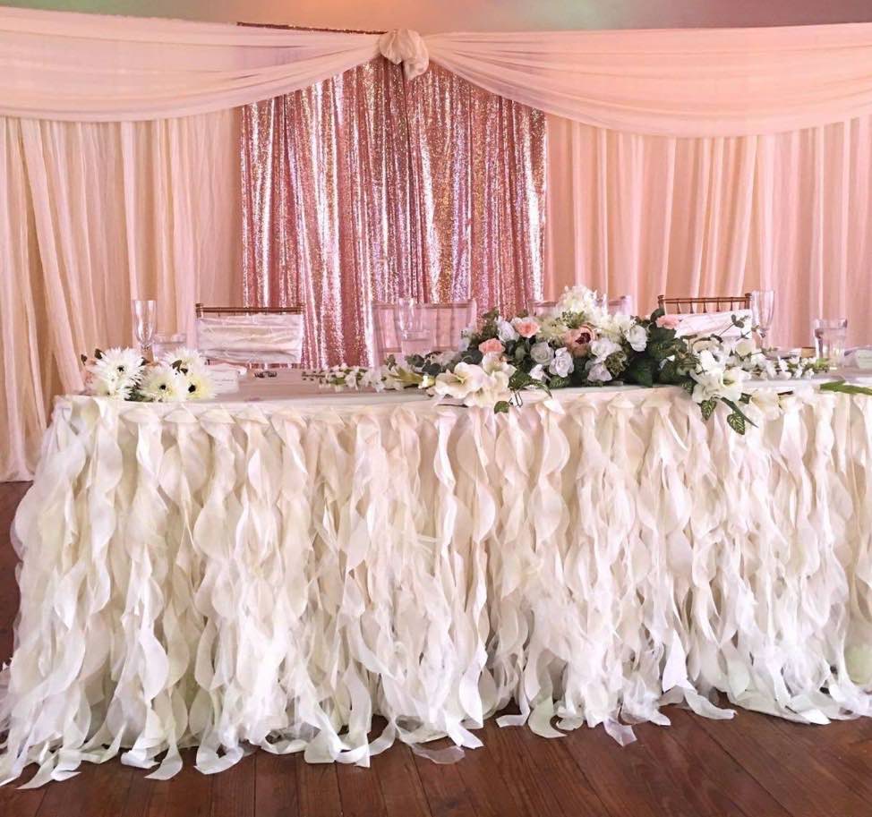 A cream ruffle head table with white and pink flowers against a rose-gold sequin backdrop.